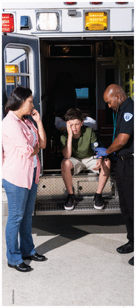 Photograph of a boy holding his head in pain, sitting out of the back of an ambulance as his mother looks on and a male paramedic takes the boy's pulse.