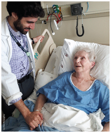 A colour photo of the author, a young man with a beard and wearing a doctor's white coat; he stands beside the hospital bed of an older female patient, wearing a blue hospital gown. They are holding hands and both are smiling..