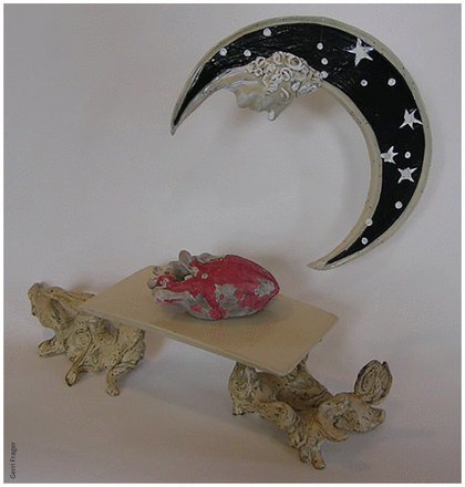 Colour photo of an artwork: a black crescent moon — with silver stars and a classically sculpted face attached to it — hangs over a small table holding a red clay heart .