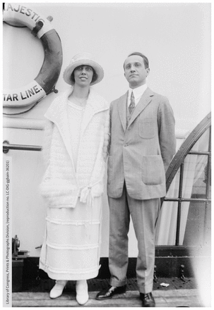 A black and white photo from the early 1900s; a man in a suit (right) stands beside a woman in a white dress and hat (left) on the deck of a ship.