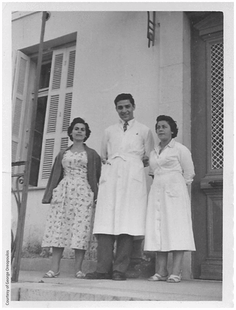 A black and white photo from the early 1960s; a young man in a white medical coat stands outside a doorway in Greece, with a woman in a dress on either side of him.