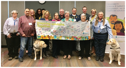 Photo of the 14 members of the Strategic Clinical Network Patient Engagement Reference Group holding a banner, and 2 dogs.